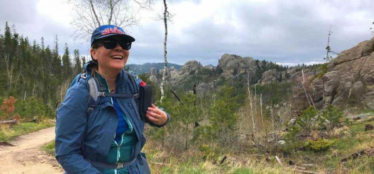 Conseils essentiels pour camper sous la pluie : restez au sec et au chaud A woman dressed in a blue rain jacket and cap, smiling and hiking on a trail surrounded by lush greenery and rocky mountains under a cloudy sky.