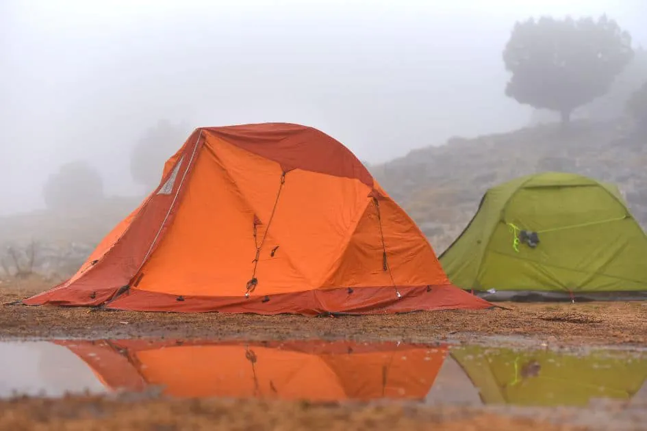 Deux tentes, une orange et une verte, installées dans un camping de montagne brumeux avec des flaques d'eau à proximité