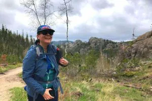 A woman dressed in a blue rain jacket and cap, smiling and hiking on a trail surrounded by lush greenery and rocky mountains under a cloudy sky.