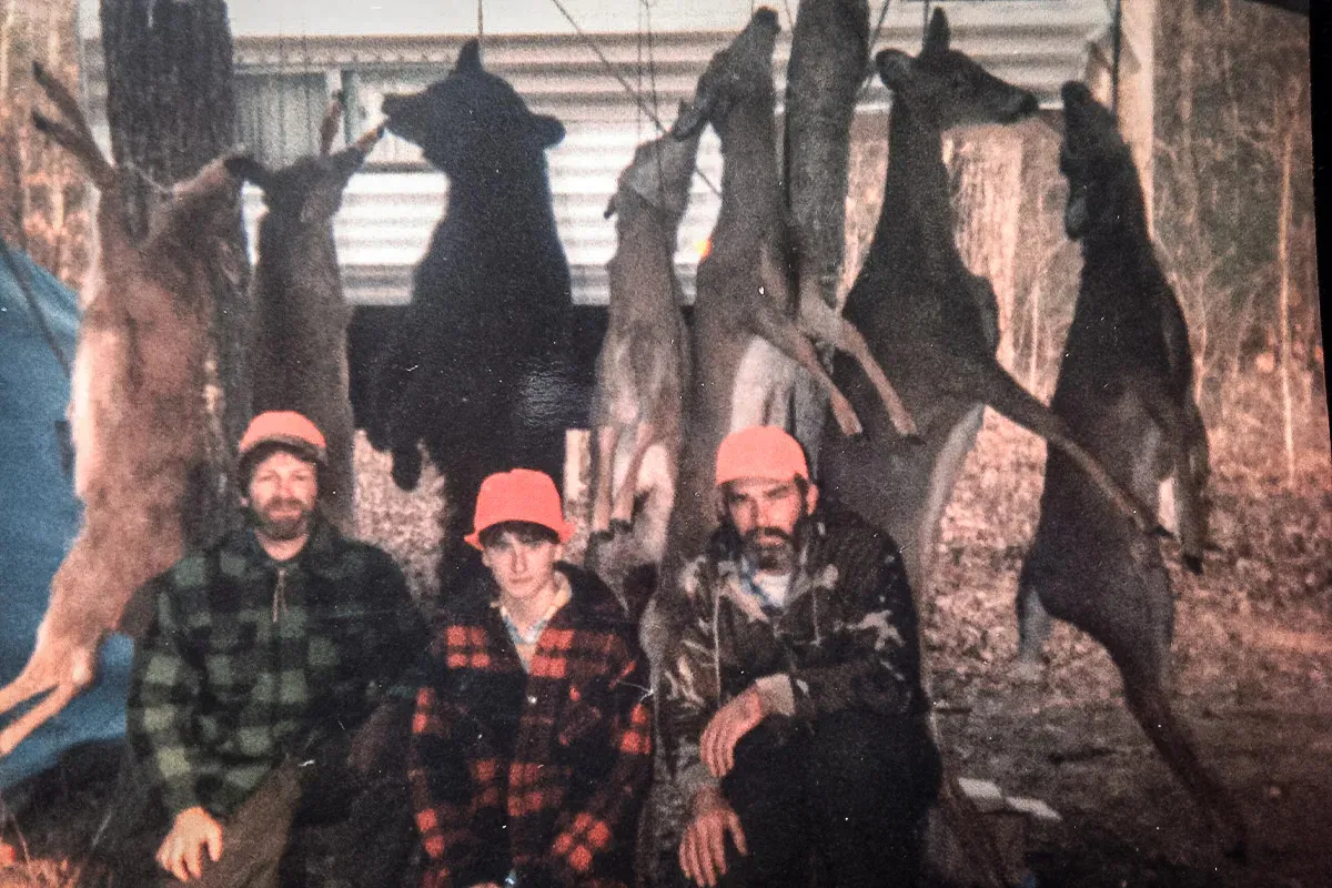 Une vieille photo de deux hommes et une jeune fille vêtue de flanelle posant devant un poteau de viande de camp de cerf