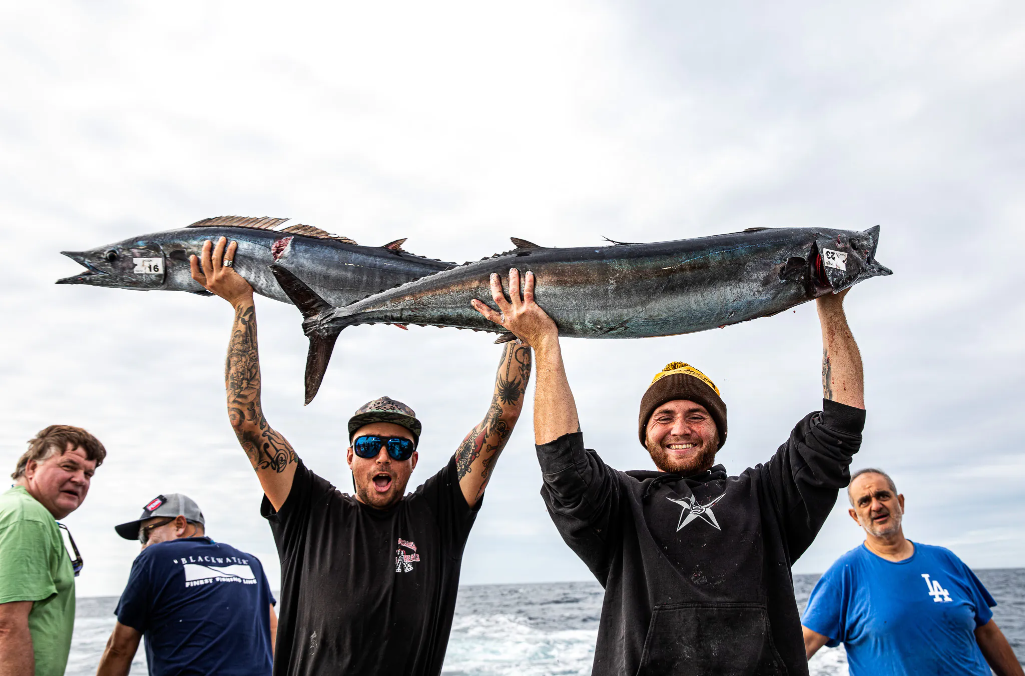 Deux gars en chemises et chapeaux noirs contiennent deux poissons wahoo au-dessus de leur tête.