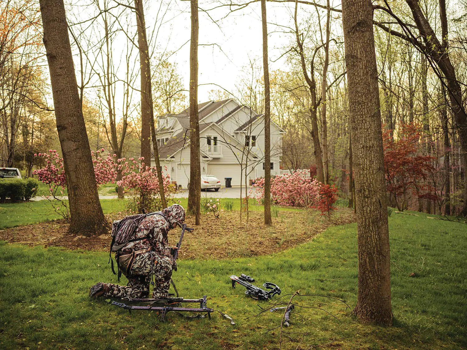 Un chasseur en camouflage complet prépare des équipements de chasse et des arbres se trouvent dans une cour arrière de banlieue.