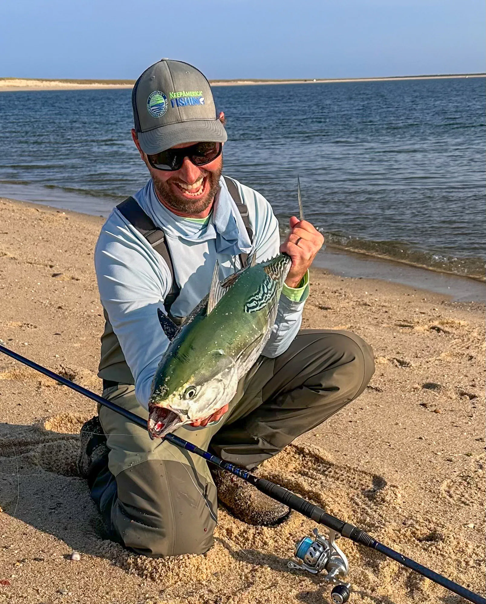 Un pêcheur dans les cuissardes s'agenouille sur le rivage sablonneux et maintient un faux albacore.