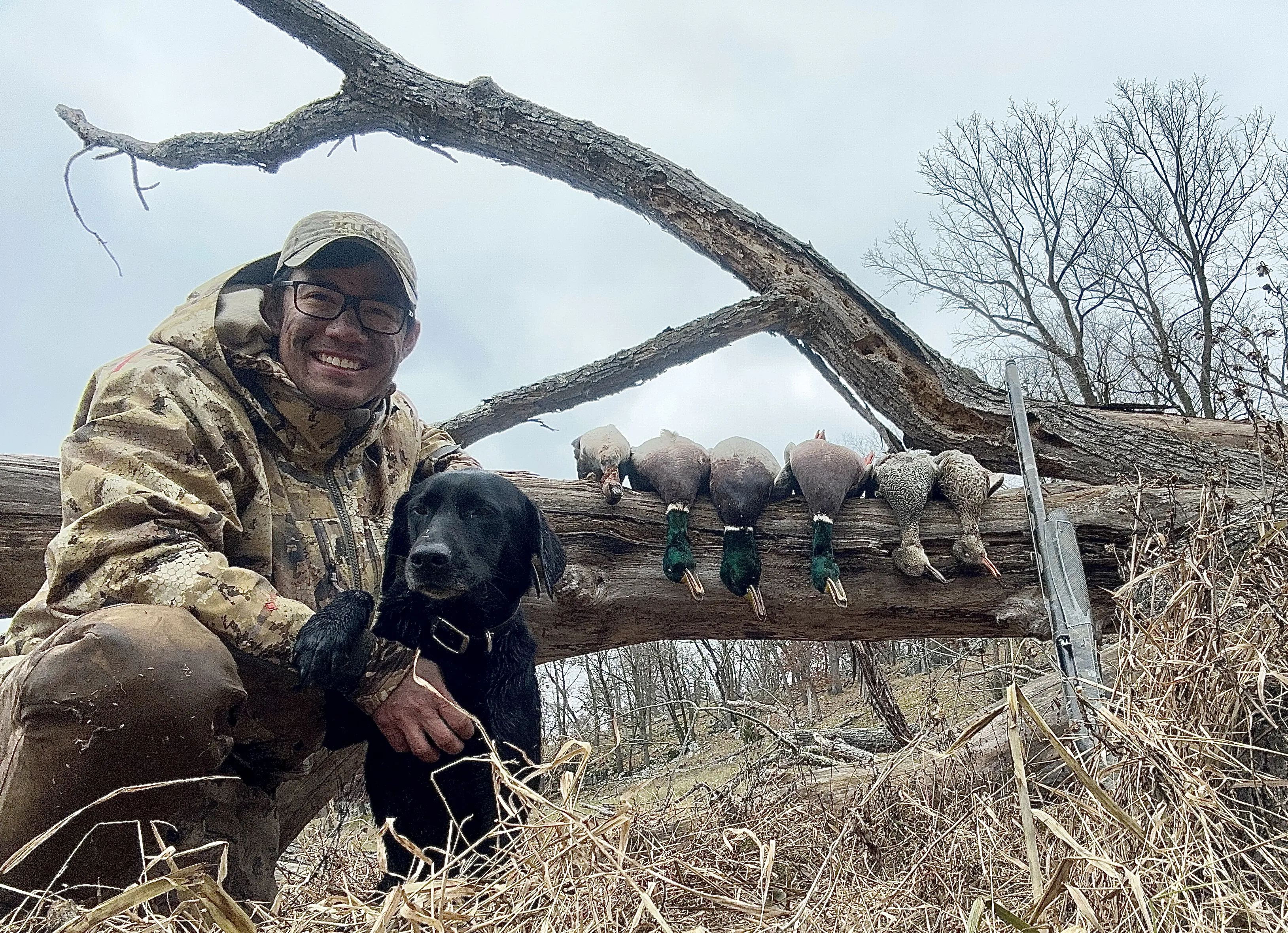Un chasseur de canards avec du maquillage camouflage est assis à côté de son laboratoire et des canards alignés sur une bûche.