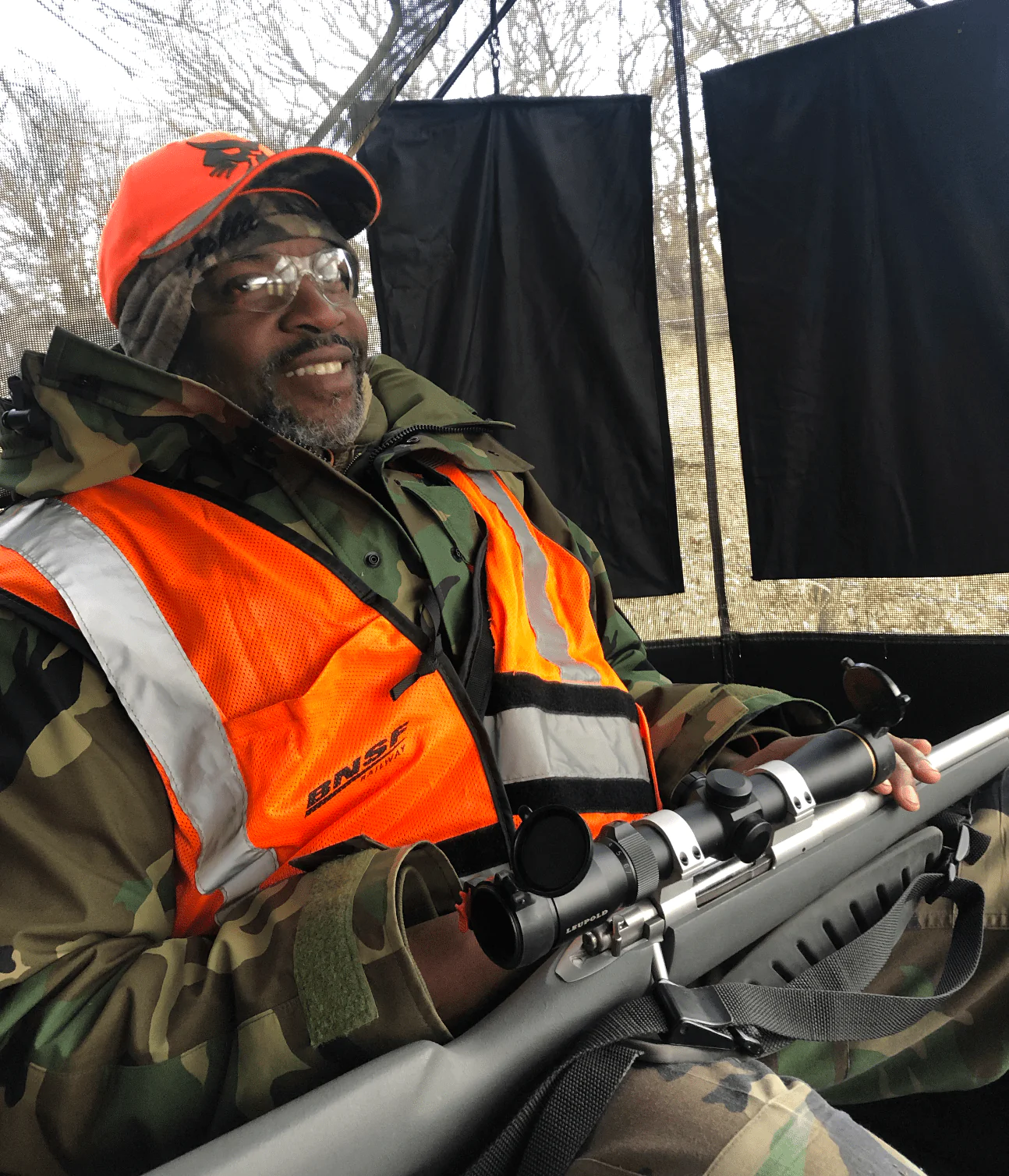 Un chasseur assis à l’intérieur d’une moustiquaire avec un camouflage, un orange flamboyant et un fusil sur ses genoux.