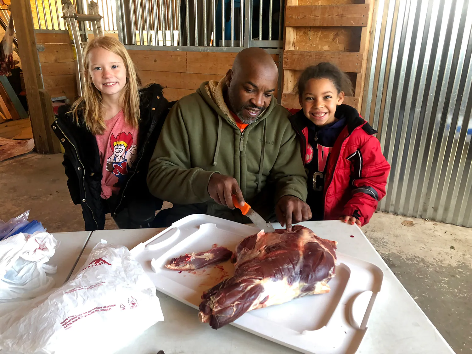 Un homme sculpte et découpe de la viande de cerf tandis que deux enfants se tiennent à ses côtés.