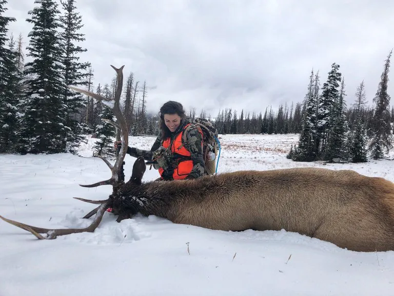 Un chasseur en kuiu et un gilet orange est assis derrière un wapiti mâle dans la neige.