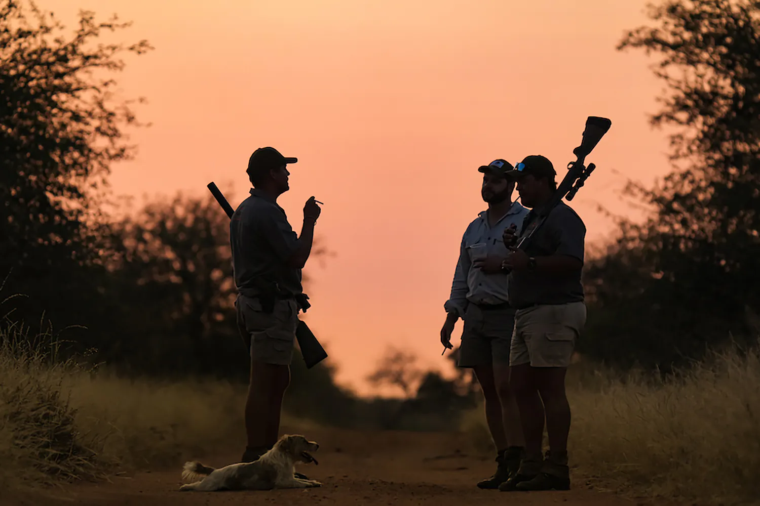trois chasseurs parlent au coucher du soleil tandis que le chien se repose aux pieds de l'un d'entre eux