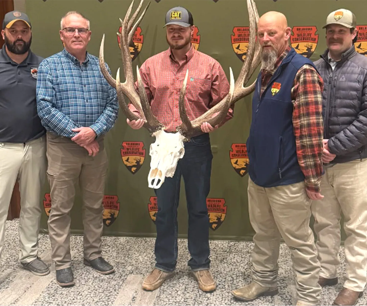 Cinq hommes se tiennent devant une marche et répètent avec le logo de conservation de la faune de l'Oklahoma et le taureau géant atypique.