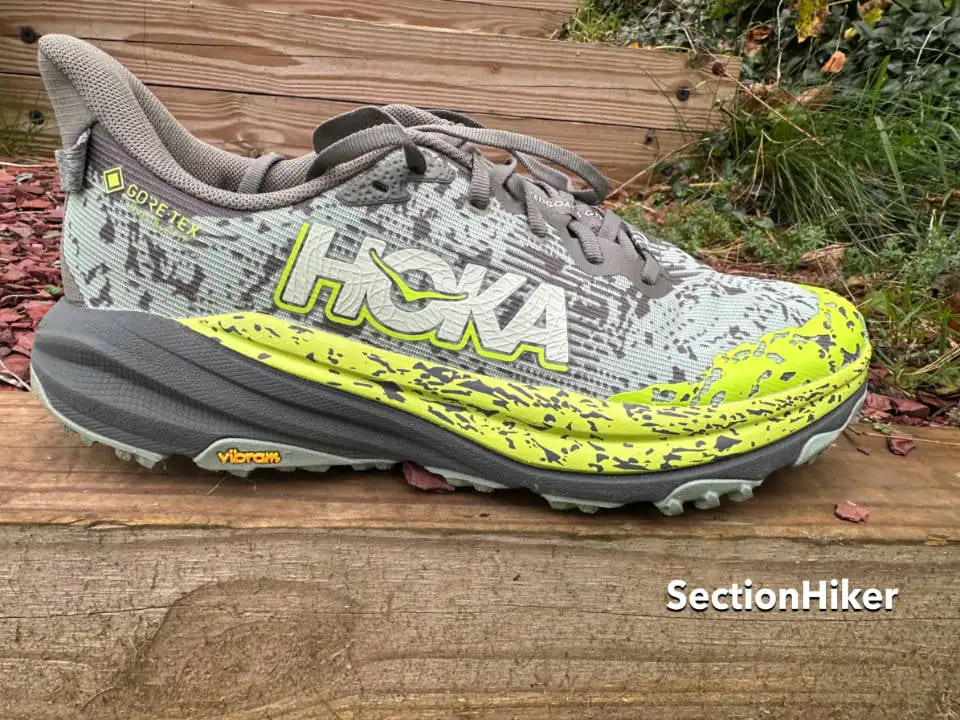 Un HOKA Maximum Cushion Trail Runner gris et vert citron avec les logos Gore-Tex et Vibram repose sur une marche en bois, entouré de paillis et d'herbe. Le mot SectionHiker apparaît dans le coin inférieur droit. Parfait pour les aventures de randonnée.